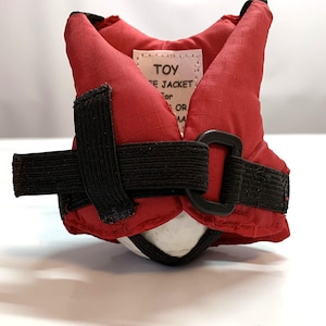 May include: Red life jacket for a toy with black straps and a label that reads "TOY LIFE JACKET for OR MA".