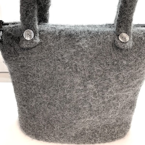 May include: A gray felt tote bag with two handles and a zipper closure. The bag features two decorative buttons on the front, near the handles. The felt material has a textured appearance.