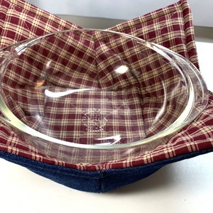 May include: An oval glass baking dish rests inside a quilted bowl cozy. The cozy features a plaid pattern in burgundy and tan, with a dark blue denim exterior. The dish is clear and has handles.