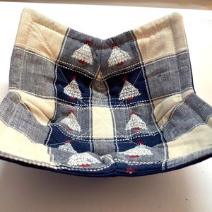 May include: A quilted bowl cozy with a plaid pattern in navy blue, cream, and gray. The cozy features embroidered Christmas trees with red accents. The bowl cozy is designed to keep food warm.