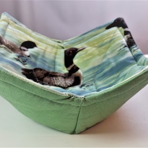 May include: A green fabric bowl with a quilted design featuring a pattern of loon birds swimming in a lake. The bowl is folded and has a soft, quilted texture.