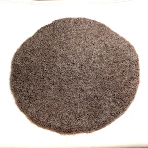 May include: A round, brown felt hat, likely a beret, sits against a white background. The hat's texture is visible, with a slightly uneven surface. The felt appears to be a solid color, with no visible patterns or embellishments.