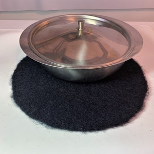 May include: A black round wool hot pad with a silver metal bowl with a lid on top.