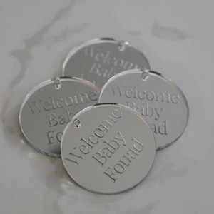 May include: Four circular silver-toned mirrored acrylic tags, each featuring a small hole at the top. The surface of each tag is engraved with the text Welcome Baby Fouad in a clean, sans-serif font, creating a reflective and smooth finish.