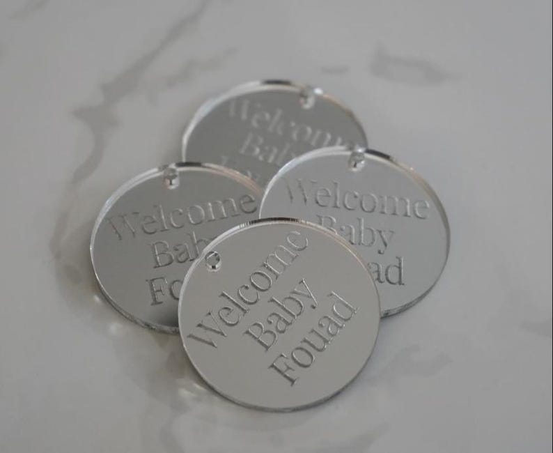 May include: Four circular silver-toned mirrored acrylic tags, each featuring a small hole at the top. The surface of each tag is engraved with the text Welcome Baby Fouad in a clean, sans-serif font, creating a reflective and smooth finish.