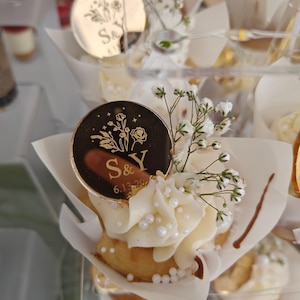 May include: A cupcake topped with white frosting, small white pearl sprinkles, and a sprig of baby's breath. A circular black acrylic topper features gold floral line art, the letters S&Y, and the date 6.13.20, all encased in a white paper liner.