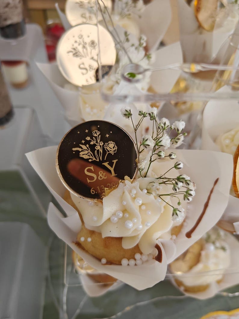 May include: A cupcake topped with white frosting, small white pearl sprinkles, and a sprig of baby's breath. A circular black acrylic topper features gold floral line art, the letters S&Y, and the date 6.13.20, all encased in a white paper liner.