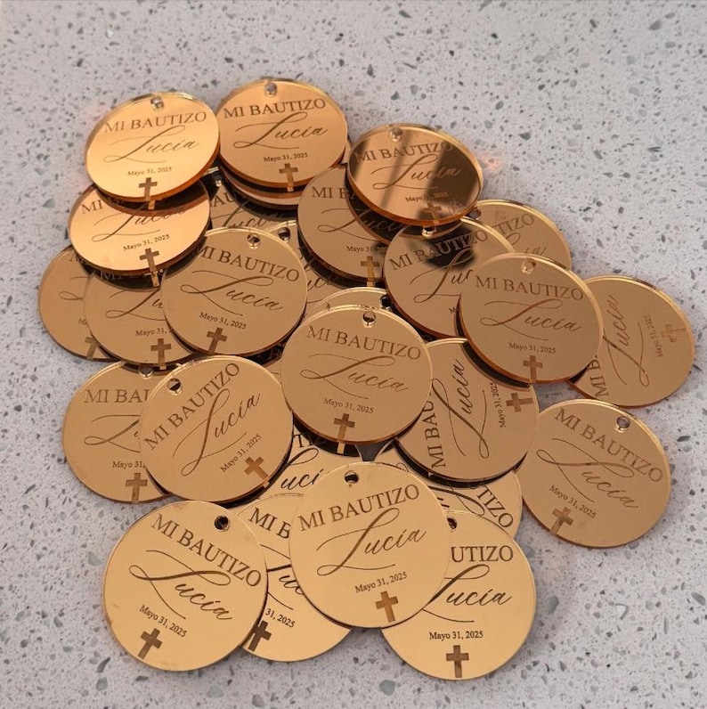 May include: A collection of circular rose gold acrylic tags, each featuring the engraved text MI BAUTIZO, the name Lucia, the date Mayo 31, 2025, and a small cross symbol. These reflective, metallic-finish discs are arranged in a scattered pile.