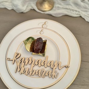 May include: A white plate with a gold rim and the words "Ramadan Mubarak" in wooden script. The plate holds a date and a decorative leaf. Two gold candle holders are in the background.