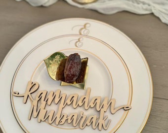 Puede incluir: Un plato blanco con borde dorado y las palabras "Ramadan Mubarak" en escritura de madera. El plato contiene un dátil y una hoja decorativa. Dos candelabros dorados están en el fondo.