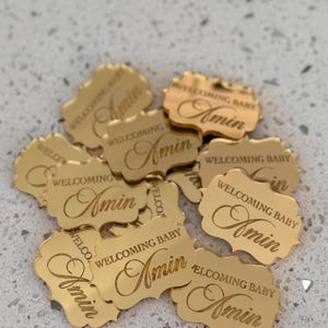 May include: A collection of small, gold-colored, reflective acrylic tags with scalloped edges. Each tag is engraved with the text 'WELCOMING BABY' in a sans-serif font and the name 'Amin' in a cursive script, arranged in a scattered pile.