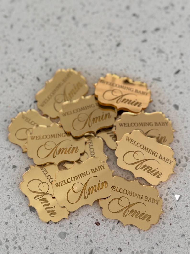 May include: A collection of small, gold-colored, reflective acrylic tags with scalloped edges. Each tag is engraved with the text 'WELCOMING BABY' in a sans-serif font and the name 'Amin' in a cursive script, arranged in a scattered pile.