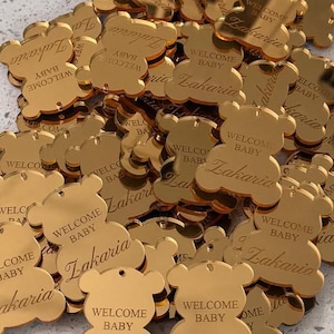 Può includere: Una collezione di etichette in acrilico color oro con finitura a specchio a forma di orsetto. Ogni etichetta presenta un piccolo foro nella parte superiore ed è incisa con il testo "WELCOME BABY" in stampatello e il nome "Zakaria" in corsivo.