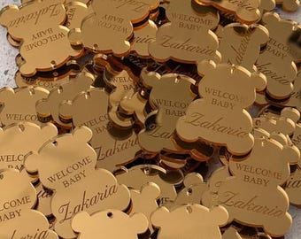 Puede incluir: Una colección de etiquetas acrílicas de color dorado con acabado de espejo en forma de ositos de peluche. Cada etiqueta tiene un pequeño orificio en la parte superior y está grabada con el texto "WELCOME BABY" y el nombre "Zakaria".