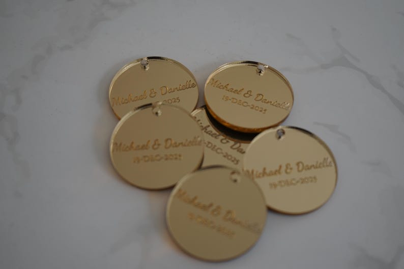 May include: A collection of five circular gold-toned acrylic tags, each featuring a small hole at the top. The tags are engraved with the text Michael & Danielle and the date 19-DEC-2025 in a cursive script font, reflecting a metallic finish.