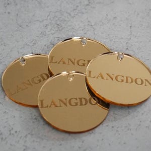 May include: Four circular gold-colored mirrored acrylic tags, each featuring the word LANGDON engraved in a serif font. Every tag includes a small pre-drilled hole at the top edge for attachment, showcasing a reflective surface and smooth edges.