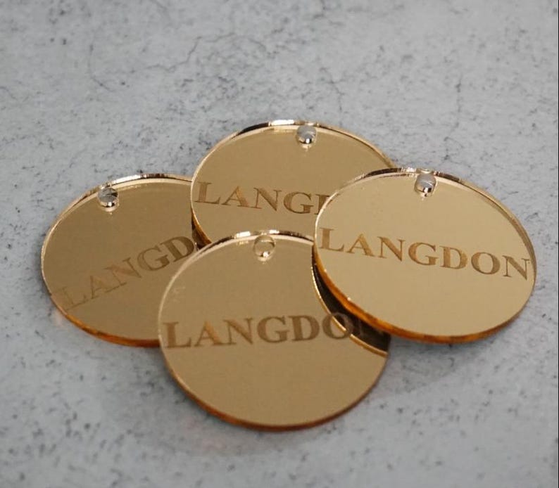 May include: Four circular gold-colored mirrored acrylic tags, each featuring the word LANGDON engraved in a serif font. Every tag includes a small pre-drilled hole at the top edge for attachment, showcasing a reflective surface and smooth edges.