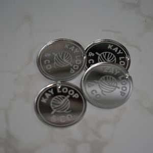 May include: Four circular silver-toned metal tags with a reflective finish. Each tag features an engraved design of a ball of yarn with a knitting needle and the text KAY LOOP & CO. arranged in a circular pattern around the central graphic.