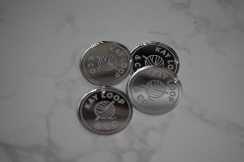 May include: Four circular silver-toned metal tags with a reflective finish. Each tag features an engraved design of a ball of yarn with a knitting needle and the text KAY LOOP & CO. arranged in a circular pattern around the central graphic.