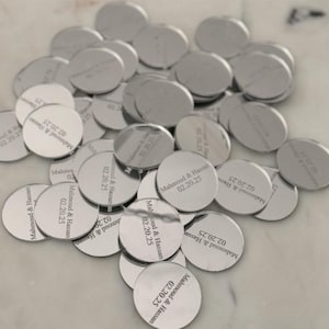 May include: A collection of circular silver-toned metal tags, each engraved with the text Mahmoud & Hassan and the date 02.20.25. The tags have a reflective, polished finish and are scattered in a pile, showing the same consistent engraving.