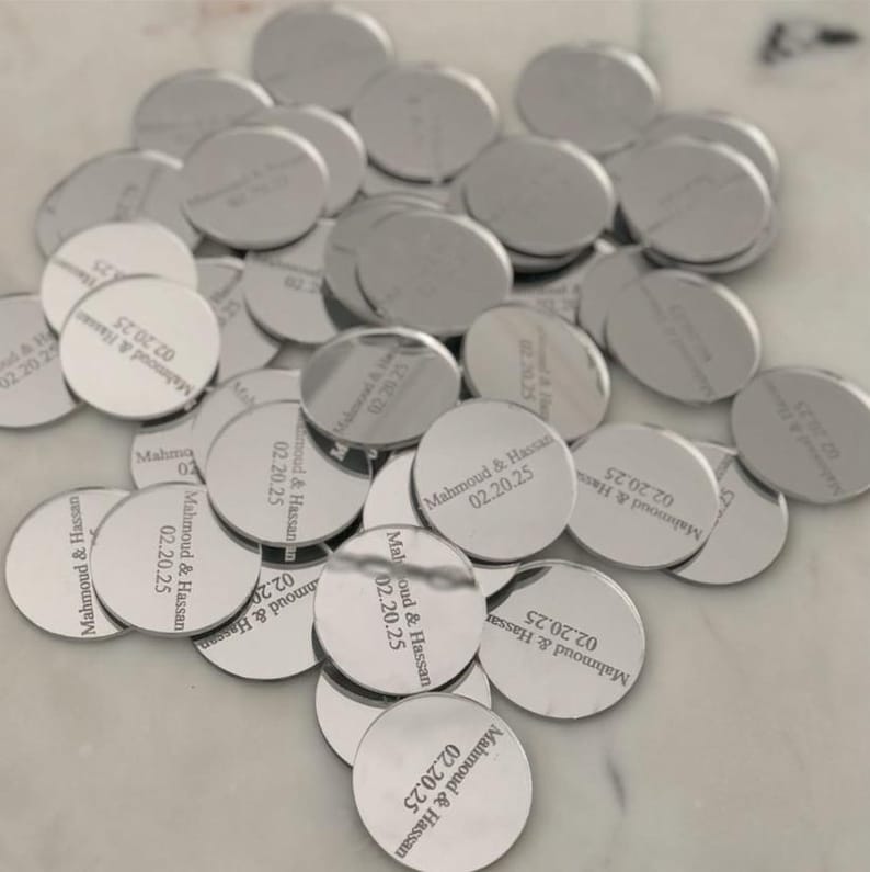 May include: A collection of circular silver-toned metal tags, each engraved with the text Mahmoud & Hassan and the date 02.20.25. The tags have a reflective, polished finish and are scattered in a pile, showing the same consistent engraving.