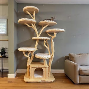 May include: A multi-level cat tree with a natural wood finish. The tree features multiple circular platforms with plush, cream-colored cushions, connected by branches. A small cat house is at the base. A gray cat is resting on one of the platforms.