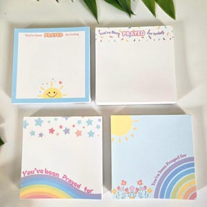 Puede incluir: Cuatro blocs de notas cuadrados con el texto "You've been Prayed for today". Cada bloc de notas presenta un diseño diferente, incluyendo un arcoíris, estrellas, un sol y bordes coloridos. Los blocs de notas son blancos con acentos pastel.