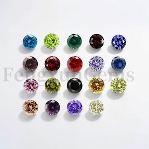 Puede incluir: Surtido de piedras preciosas redondas y facetadas en varios colores, incluyendo azul, verde, rojo, morado, negro y amarillo. Las piedras preciosas están dispuestas en filas sobre una superficie blanca.