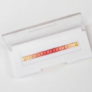 May include: A white plastic case with a clear lid, open to reveal a row of rectangular gemstones. The gemstones transition in color from yellow to red. The case is on a white background.