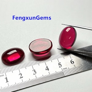 以下が含まれることがあります： 3つの楕円形に磨かれた赤い宝石。1つの宝石はピンセットで持たれています。ミリメートル単位の目盛りが付いた定規がフレーム内にあります。「FengxunGems」というテキストが左上にあります。