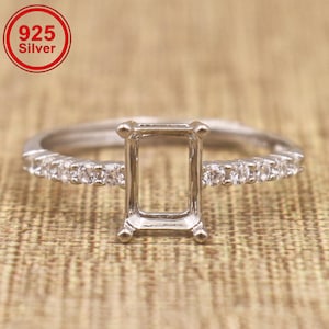 May include: A 925 silver ring setting featuring a rectangular prong mount at the center, flanked by a row of small clear stones along each side of the band. The metal has a polished finish and the design is intended for a center gemstone.