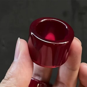 Puede incluir: Un anillo cilíndrico de color rojo intenso con un acabado pulido y brillante. El anillo se sostiene sobre un fondo oscuro, mostrando su cualidad translúcida y su superficie lisa. El anillo tiene una banda ancha y un centro hueco.