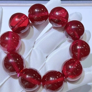Puede incluir: Una pulsera con grandes cuentas redondas rojas translúcidas. Las cuentas tienen estrías internas y están ensartadas en un cordón rosa. La pulsera se muestra sobre un fondo blanco, resaltando el color vibrante y la claridad de las cuentas.