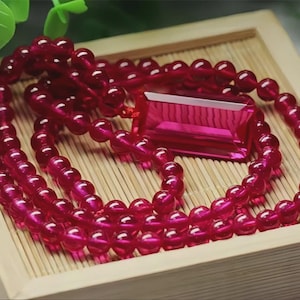 Puede incluir: Un vibrante collar de cuentas rojas con un colgante rectangular. El collar está compuesto por pequeñas cuentas redondas y un gran colgante facetado. La joya se exhibe sobre una superficie de madera clara.