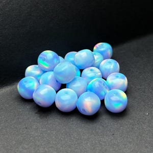 May include: A cluster of light blue opal beads, each round and displaying an iridescent sheen. The beads show a mix of colours, including blue, purple, and green. The beads are smooth and polished, with a subtle glow.