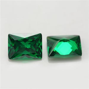 May include: Two emerald green rectangular cut gemstones. The gemstones are faceted and have a high polish.