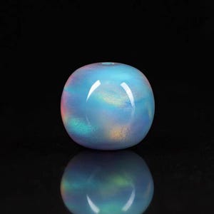 Puede incluir: Una esfera de vidrio iridiscente con una pequeña abertura en la parte superior. La esfera tiene una combinación de colores azul y morado con toques de verde y amarillo. La superficie es lisa y reflectante, con un reflejo espejado debajo.