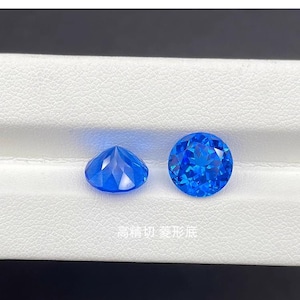 Puede incluir: Dos gemas azules facetadas, redondas y de talla brillante. Una se muestra de lado, revelando su base puntiaguda, y la otra se ve desde arriba. Las gemas se exhiben sobre una superficie blanca. Se ve texto en caracteres chinos.