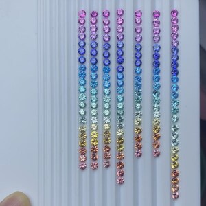 Op de afbeelding: Rijen kleine, ronde edelstenen in een regenboog van kleuren, variërend van roze en paars tot blauw, groen, geel en oranje. De edelstenen zijn in verticale lijnen op een wit oppervlak gerangschikt.
