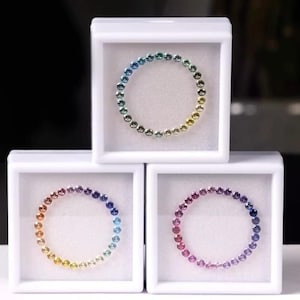 Puede incluir: Tres cajas de exhibición blancas contienen cada una un anillo de pequeñas piedras preciosas de colores. Los anillos muestran un degradado de colores, incluyendo azul, verde, amarillo, naranja y rosa. Las piedras preciosas están sobre un fondo blanco.