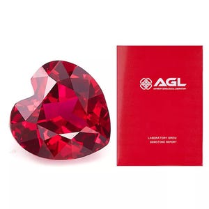 Puede incluir: Una piedra preciosa de rubí de laboratorio en forma de corazón, de color rojo. La piedra preciosa está acompañada de una tarjeta roja con el texto "AGL Antwerp Gemological Laboratory" y "Laboratory Grown Gemstone Report".