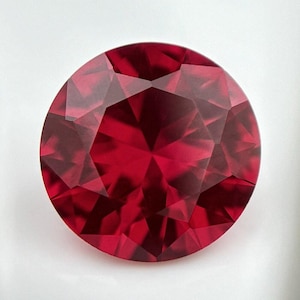 May include: A deep red gemstone with a round cut and many facets. The gemstone is set against a white background.