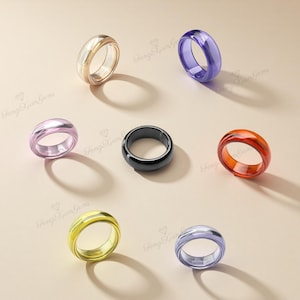 May include: A collection of colorful rings in various shades, including gold, purple, pink, black, and orange. The rings are displayed on a light beige surface, with shadows adding depth. The rings have a smooth, polished appearance.