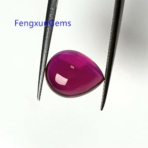 Puede incluir: Una piedra preciosa de color rosa brillante en forma de lágrima, sostenida por unas pinzas. La piedra preciosa es lisa y pulida.