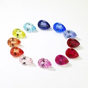 May include: A collection of teardrop-shaped gemstones in a rainbow of colors, including yellow, orange, red, pink, purple, blue, and green. The gems are arranged in a circular pattern against a white background.