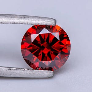 May include: A round, red gemstone with a faceted cut. The gemstone is held in place by a pair of tweezers.
