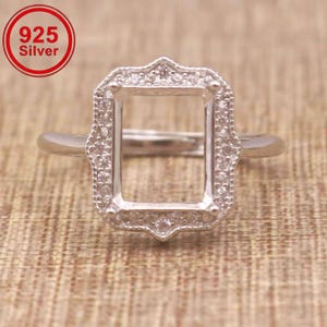 May include: A 925 silver ring setting featuring a rectangular centre mount surrounded by small clear stones. The metal band is smooth and polished, and the rectangular frame has a decorative scalloped edge with embedded stones. Text 925 Silver.