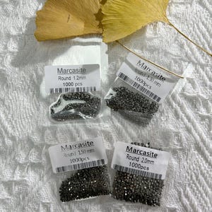 Pode incluir: Quatro sacos de plástico transparente contendo pequenas pedras de marcassita redondas cinza-escuras. Cada saco tem uma etiqueta branca com o texto Marcasite, o tamanho em milímetros e 1000 pcs. Os tamanhos são 1,2mm, 1,30mm, 1,50mm e 2,0mm.