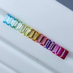 Könnte beinhalten: Eine Reihe rechteckiger Edelsteine in Regenbogenfarben, von Hellblau bis Dunkelmagenta. Die Edelsteine sind linear angeordnet und zeigen einen Farbverlauf.