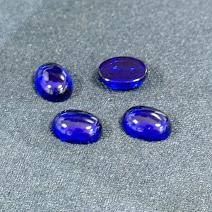 Op de afbeelding: Vier ovaalvormige, diepblauwe cabochon edelstenen met een glad, gepolijst oppervlak. Elke steen heeft een afgeronde, koepelvormige bovenkant en een platte basis, die het licht reflecteert met een glanzende glans. De stenen zijn in een losse groep gerangschikt.